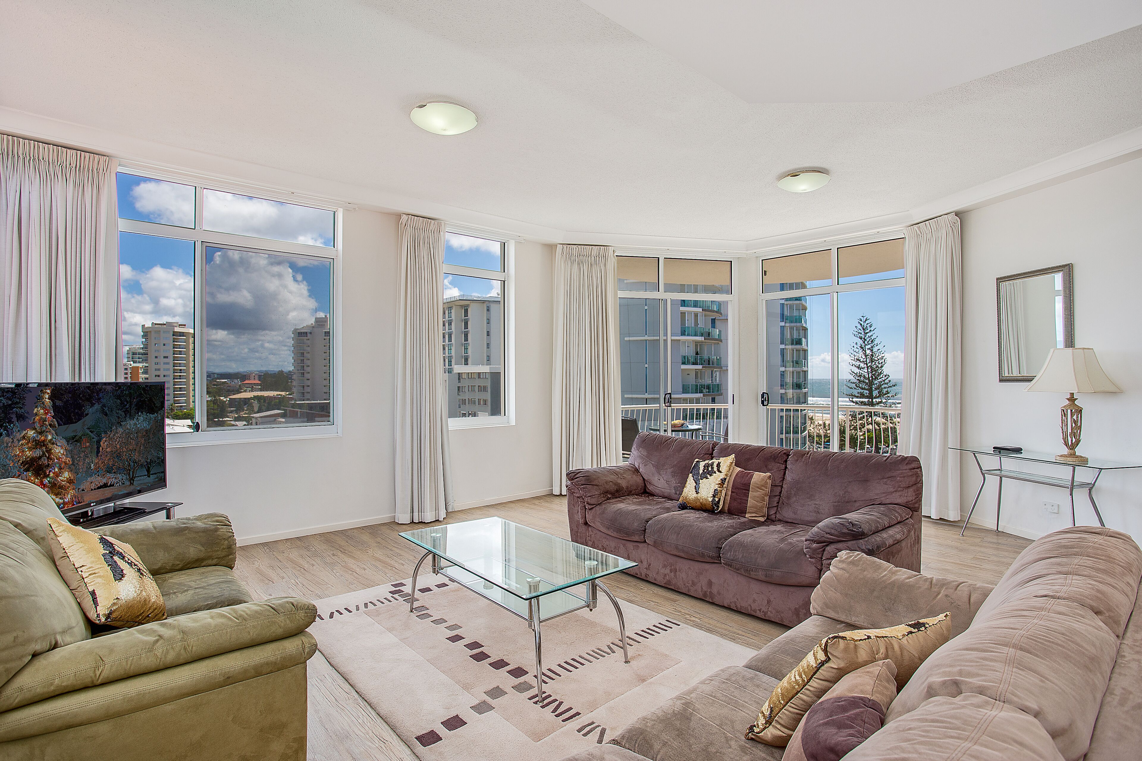 A modern living room with a glass coffee table, two sofas, a TV, and large windows offering a city view.