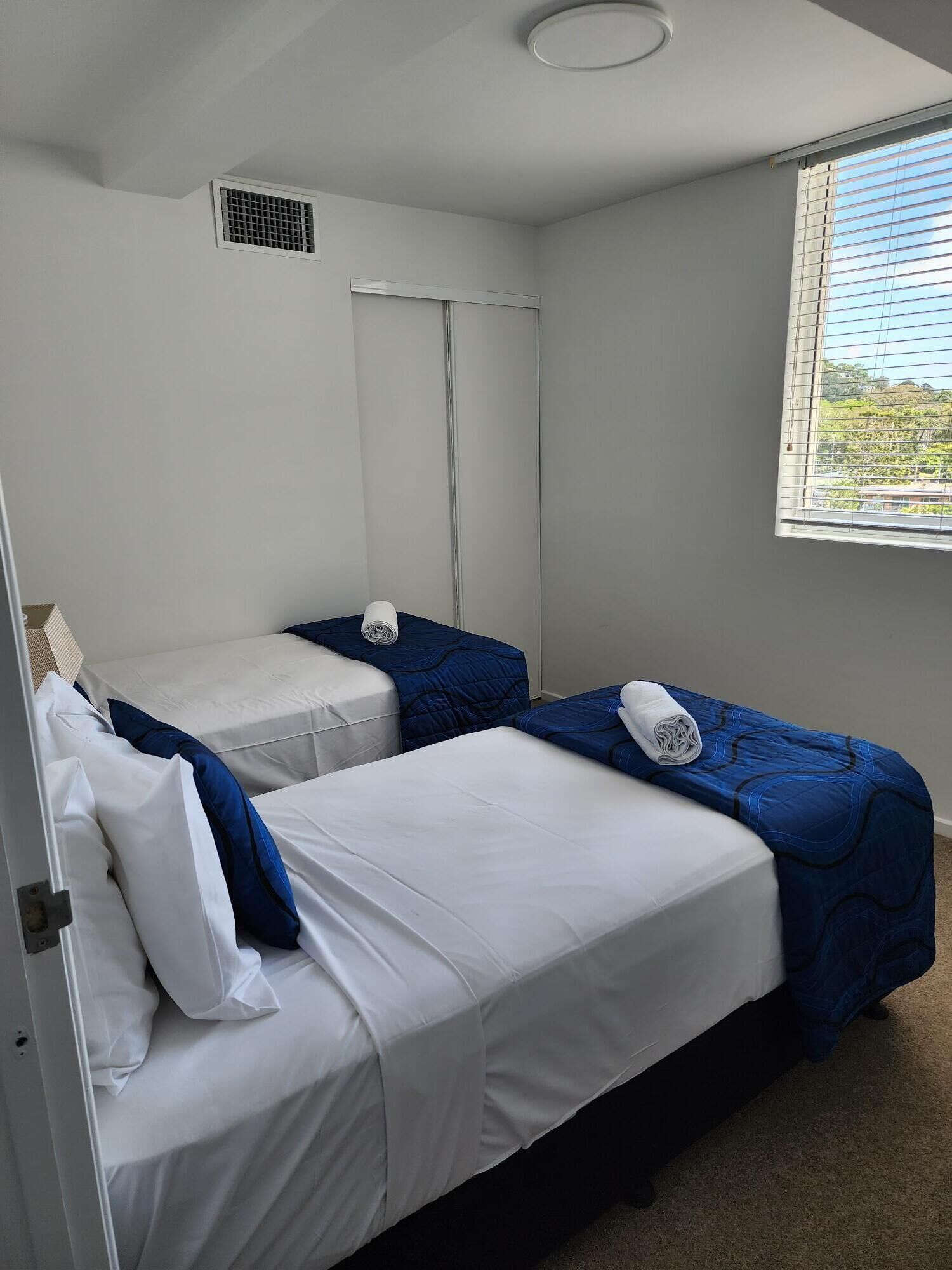 A room with two beds, a window with blinds, a door, and a ceiling vent.
