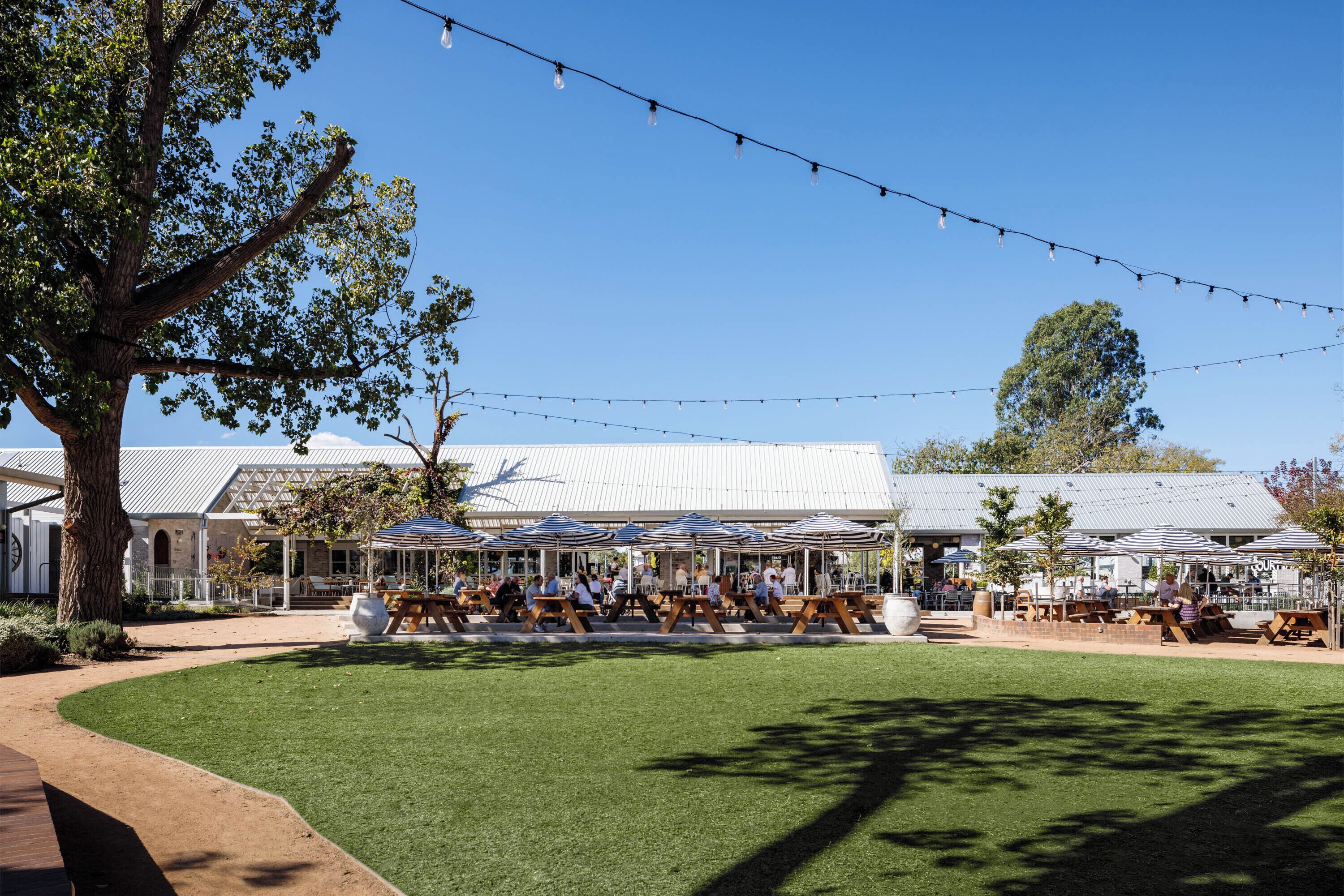 An outdoor seating area with tables and umbrellas, surrounded by trees and a building with a metal roof.