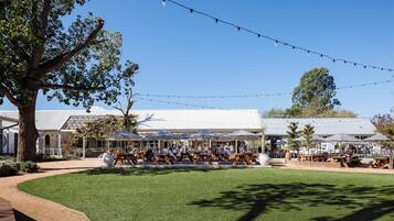 An outdoor seating area with tables and umbrellas, surrounded by trees and a building with a metal roof.