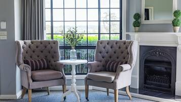 Une pièce comprenant deux fauteuils rembourrés, une petite table ronde, une cheminée et une grande fenêtre avec des rideaux.
