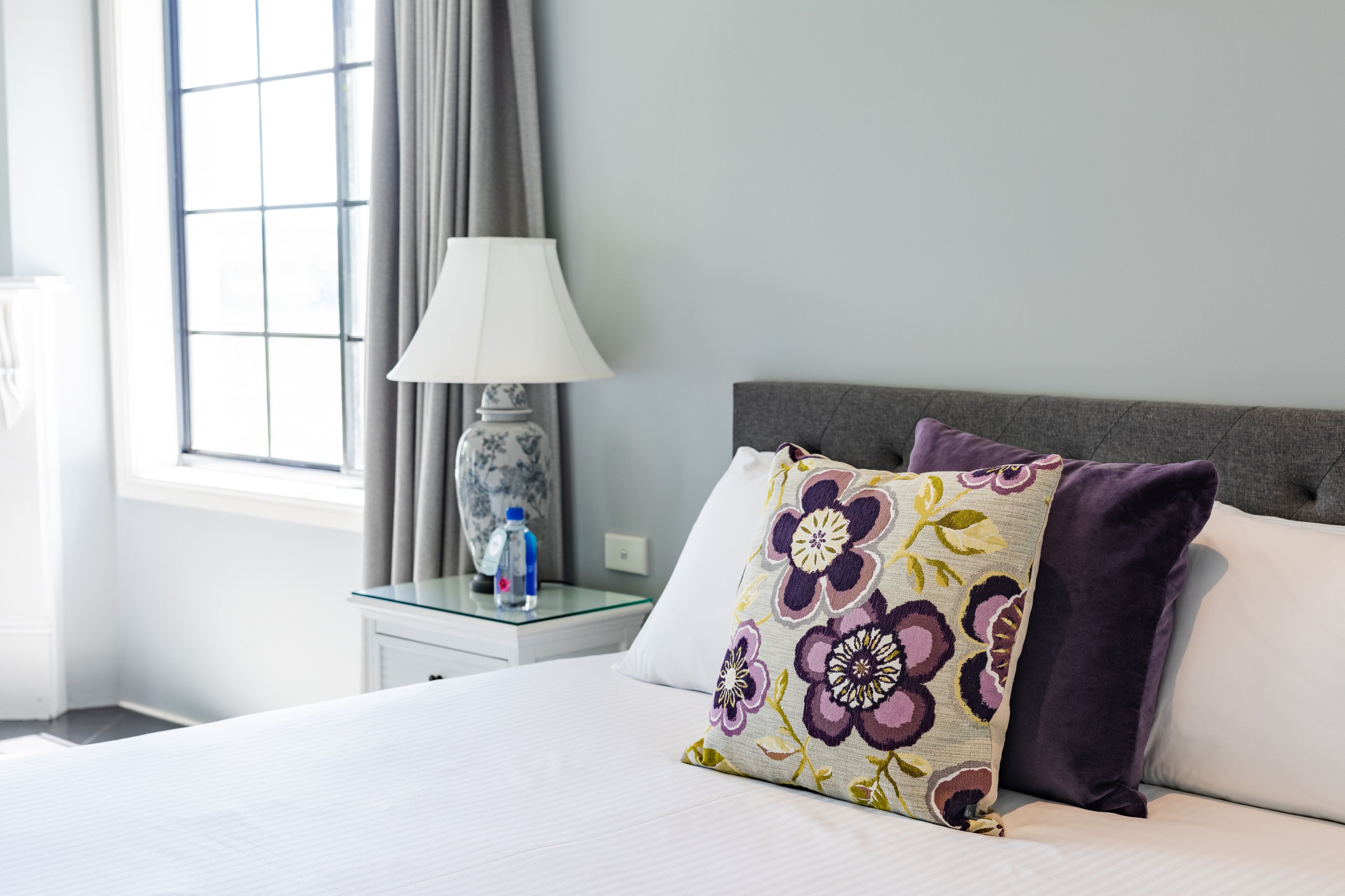 A neatly made bed with floral pillows, a bedside table with a lamp and a bottle, and a window with curtains.