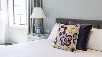 A neatly made bed with floral pillows, a bedside table with a lamp and a bottle, and a window with curtains.