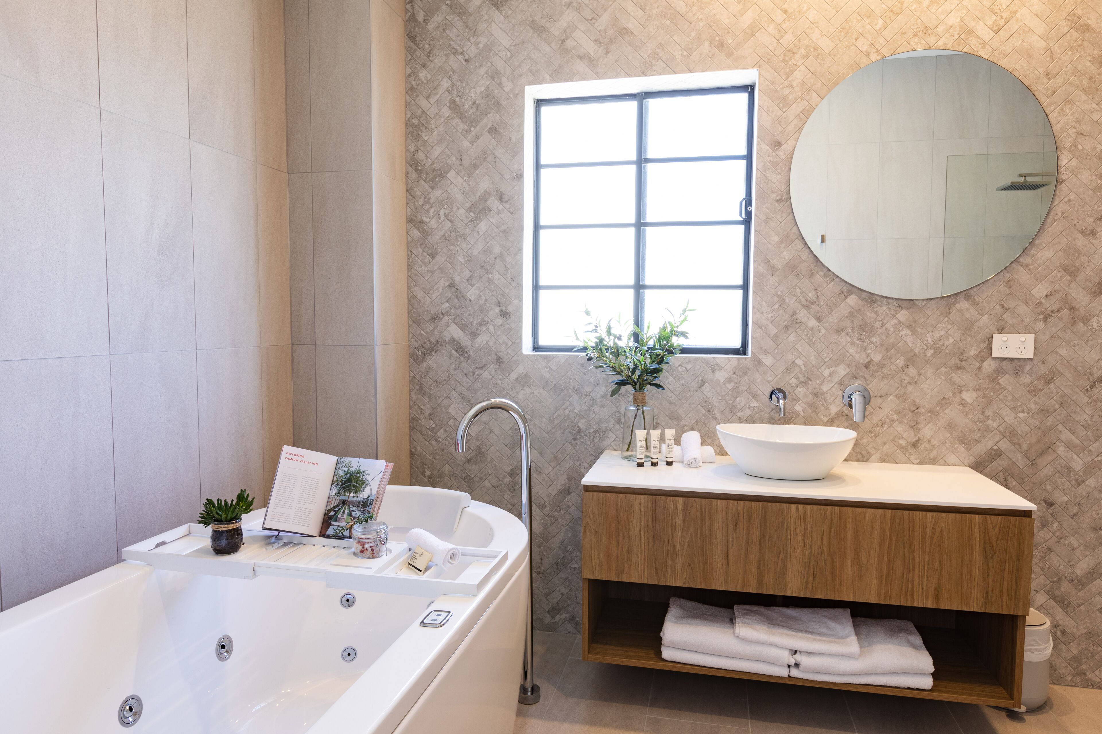 Un bany modern amb una banyera autònoma, un mirall rodó i un penjador de fusta amb un lavabo blanc.