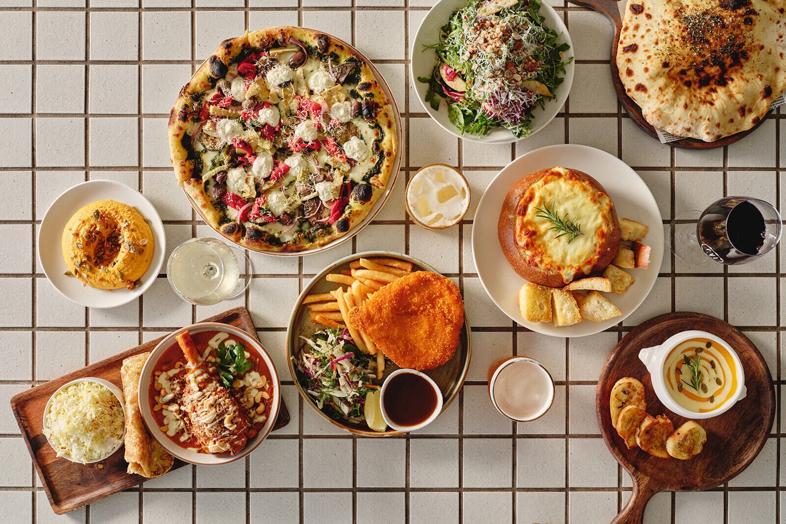A variety of dishes including pizza, salad, and bread, arranged on a tiled table.