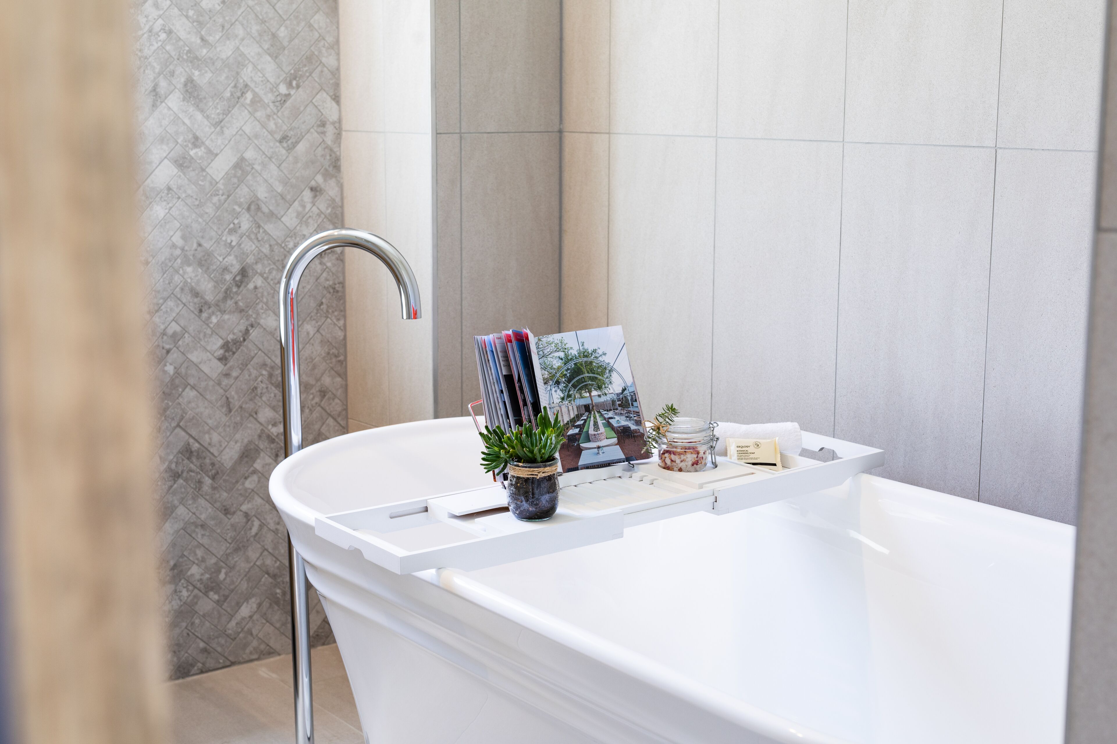 Un baño moderno con una bañera independiente, una planta en maceta, una revista y un pequeño tarro sobre el borde.