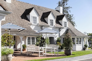 A building with a white facade, multiple windows, and a brick chimney. There are outdoor seating areas with umbrellas and potted plants.
