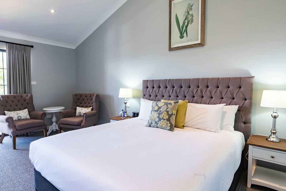 A hotel room with a large bed, two armchairs, a round table, and a framed botanical illustration on the wall.