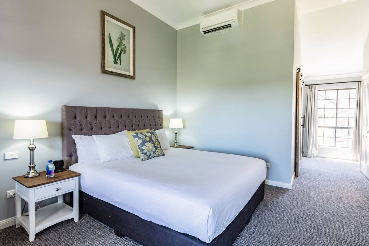 A neatly made bed with a tufted headboard, two bedside tables with lamps, a framed botanical print on the wall, and an air conditioning unit.