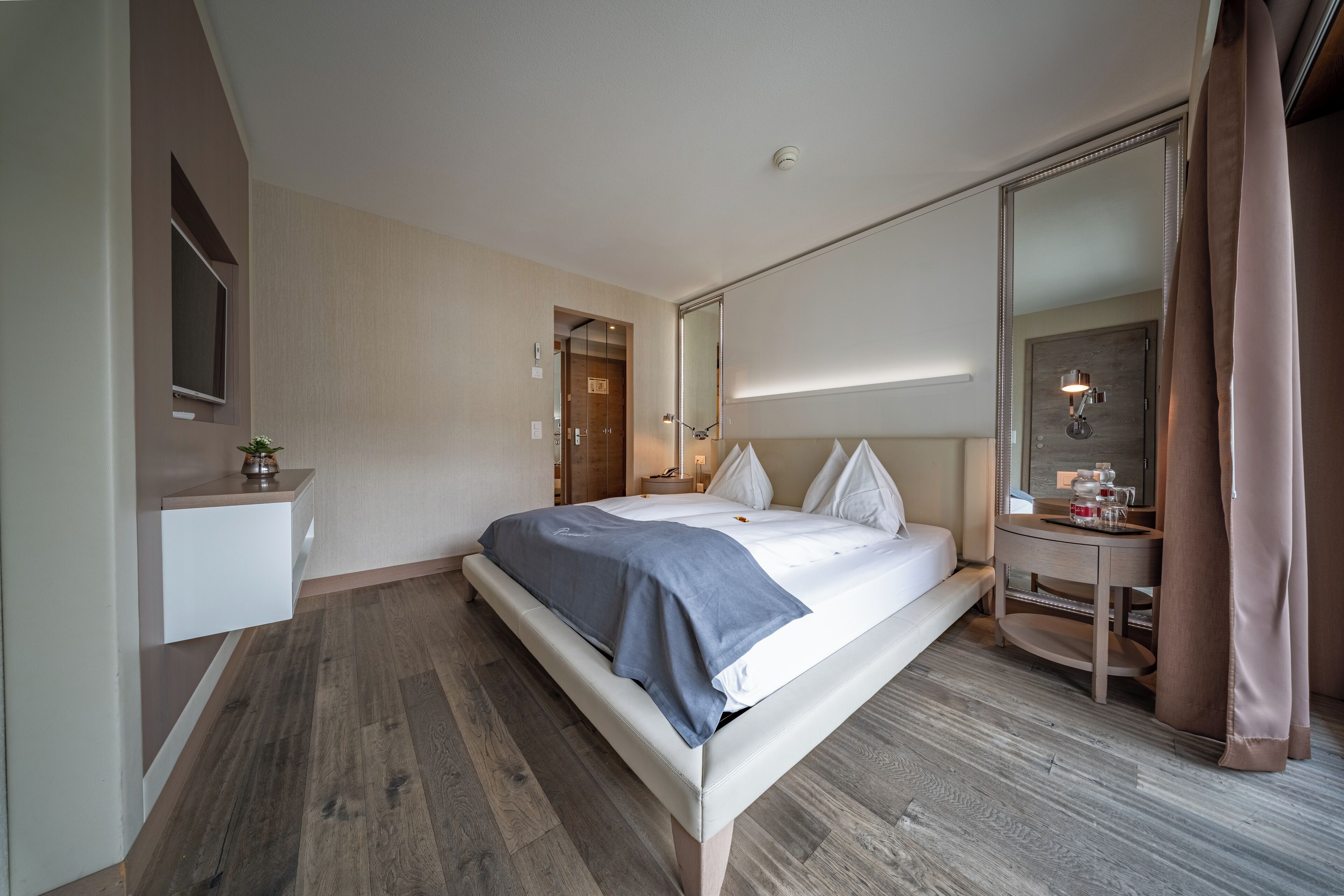 A modern hotel room with a large bed, a bedside table, a mirror, and a small desk with a lamp.