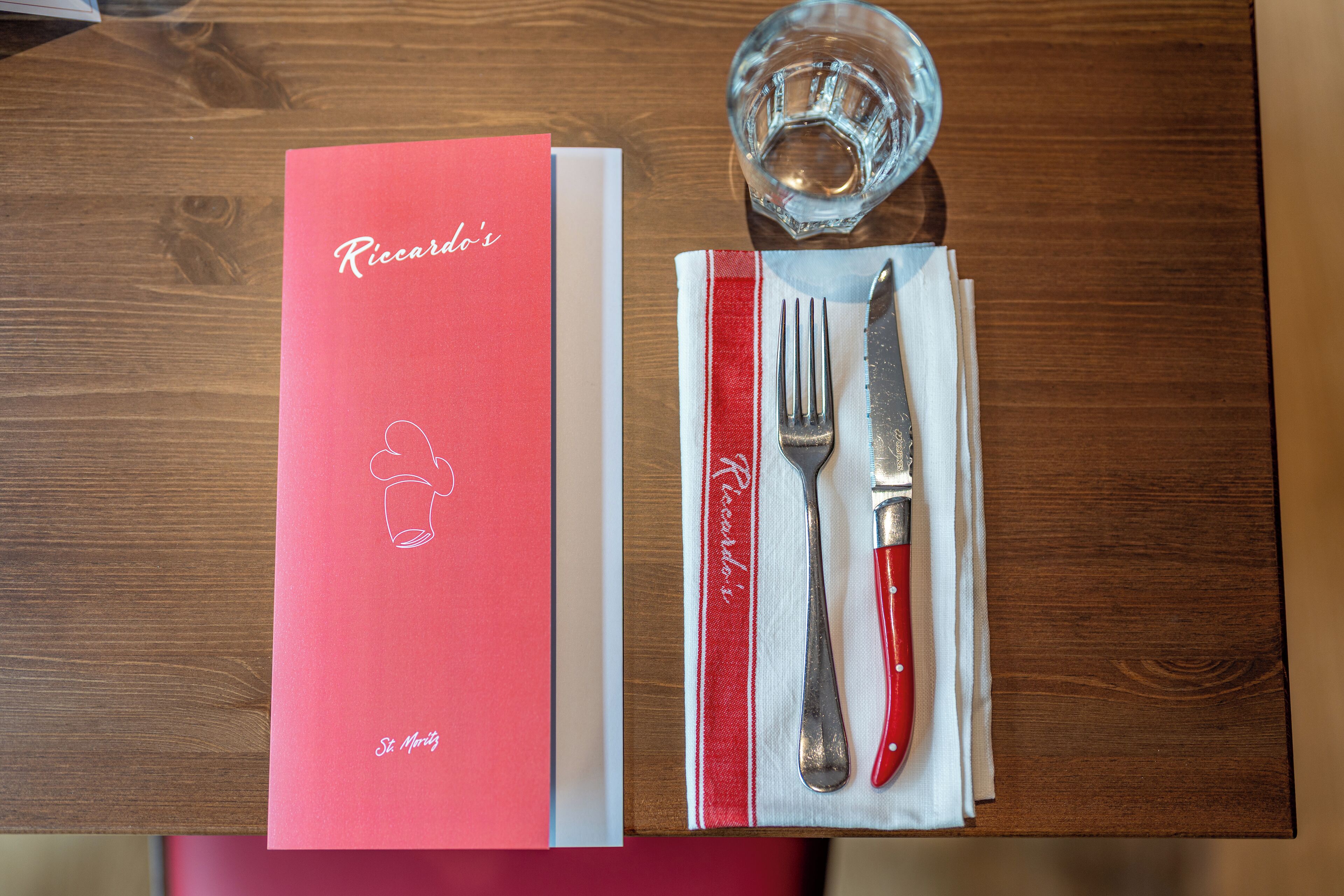 A table with a red menu, a glass of water, and cutlery on a napkin.