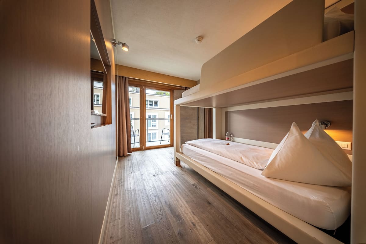 A modern hotel room with a wooden floor, a bed with white bedding, and a view of buildings through large windows.