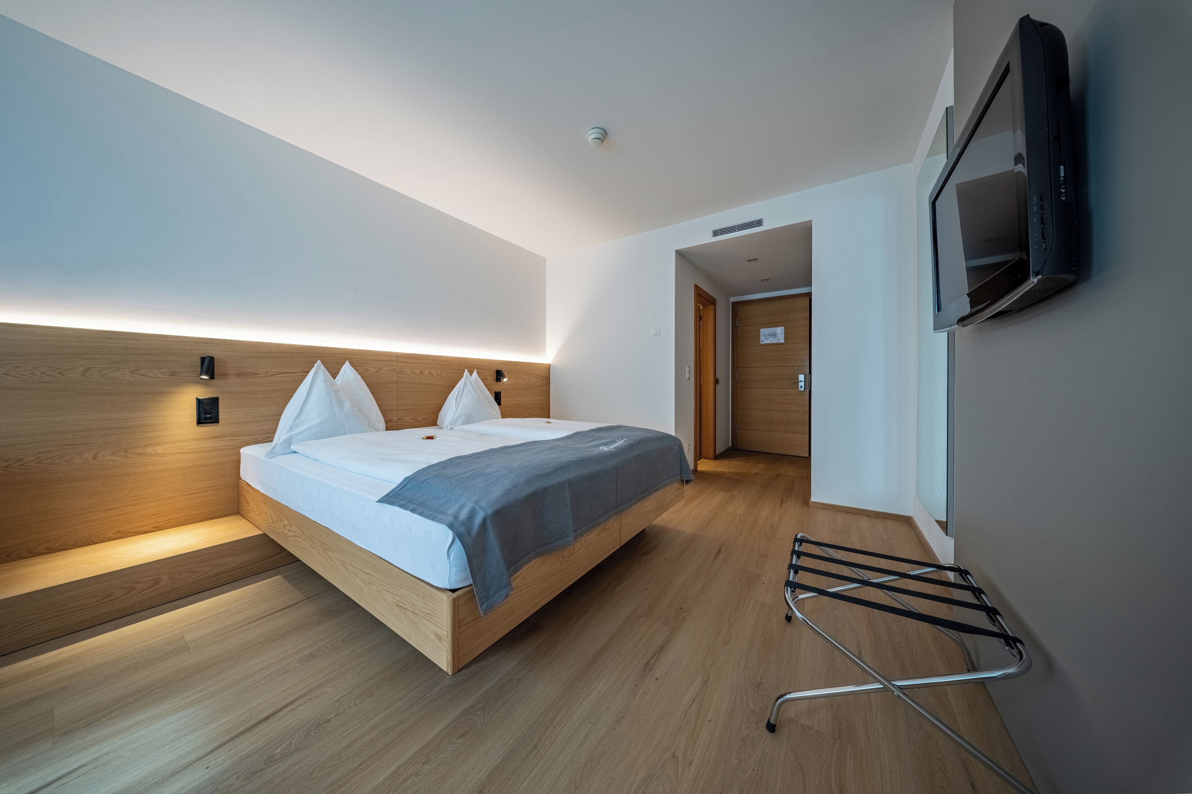 A modern hotel room with a wooden bed, a flat-screen TV mounted on the wall, and a built-in bench at the foot of the bed.