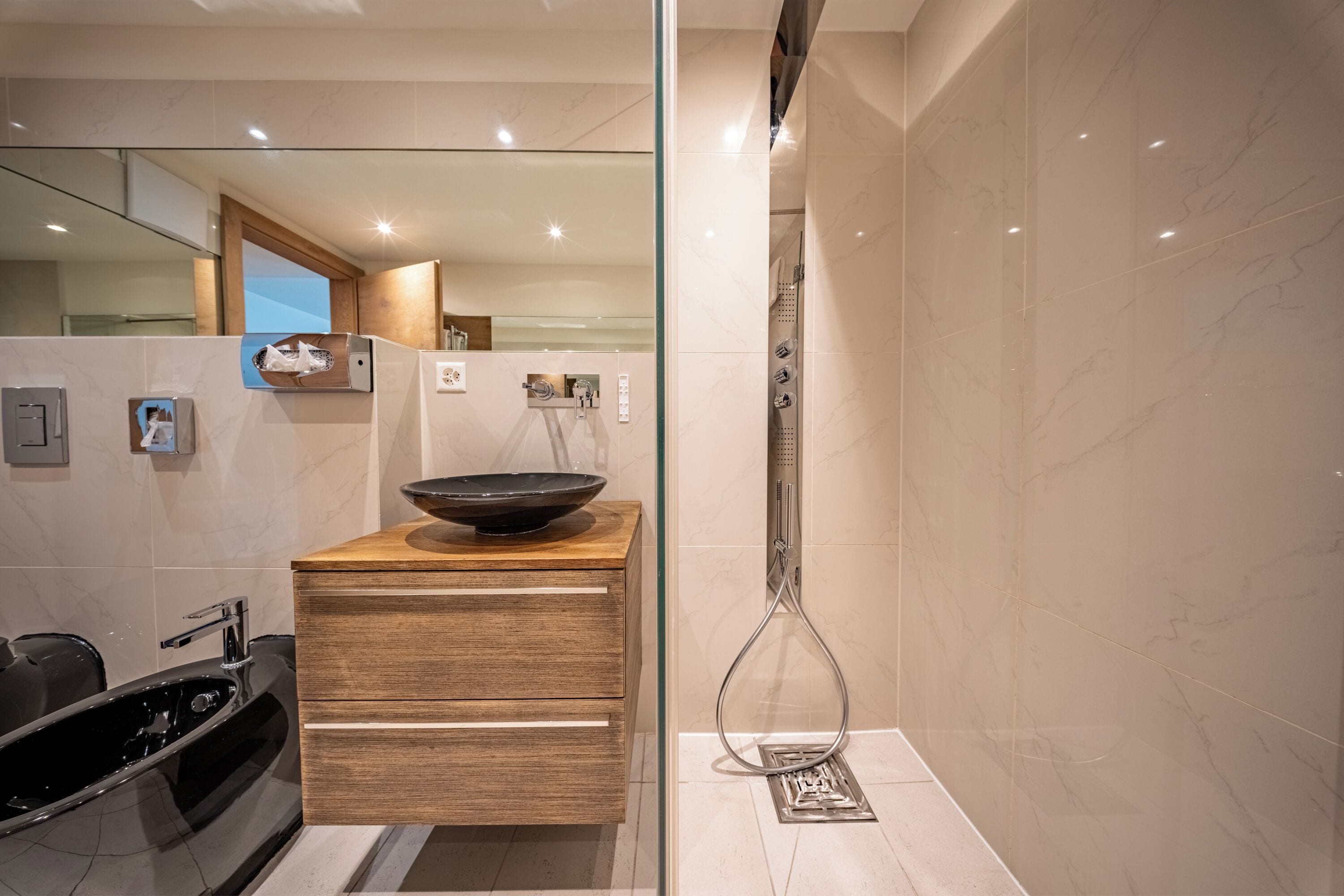 Une salle de bain moderne avec un meuble-lavabo en bois, un lavabo noir, une cabine de douche en verre et une zone de douche carrelée en marbre.