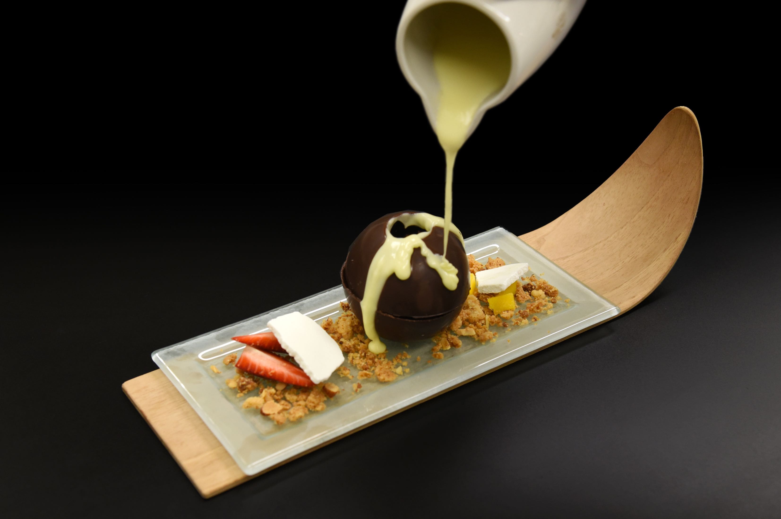 Ein Dessert mit einer Schokoladensphäre, einer weißen Soße und einem Holzlöffel auf einem rechteckigen Teller.