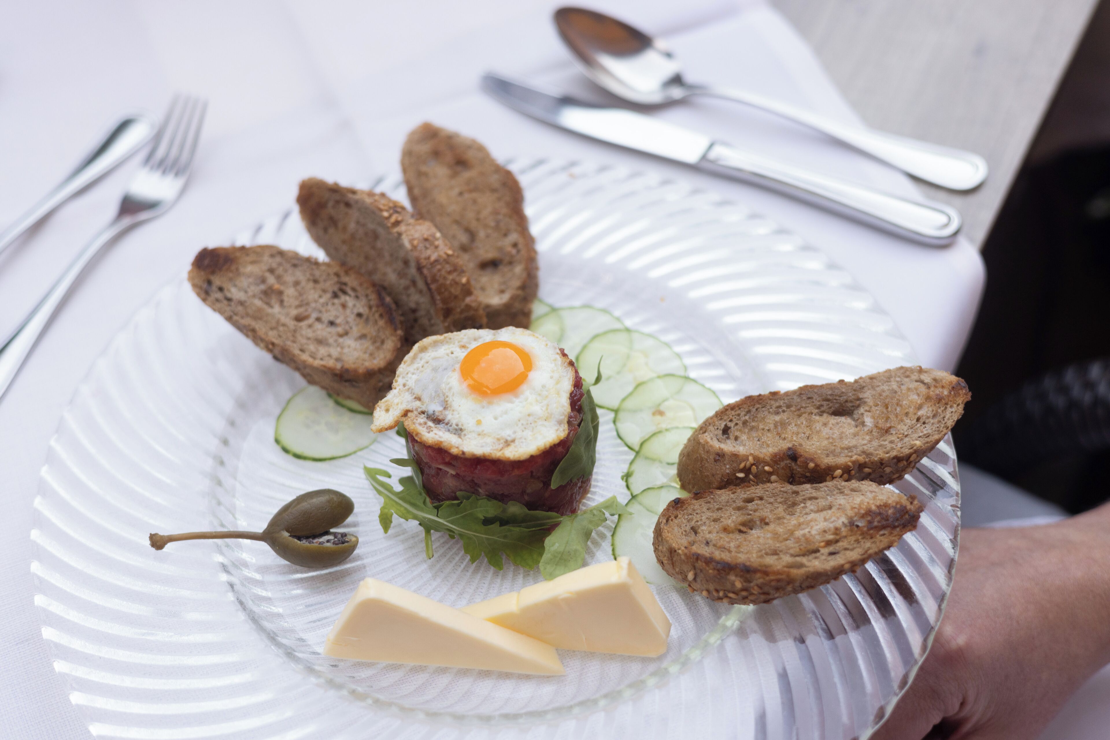 A plate with a steak tartare, cheese, cucumber slices, and bread.