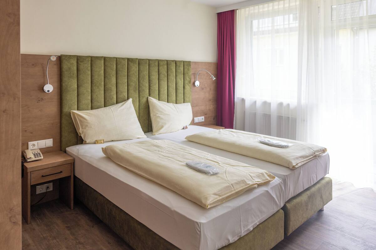 A double bed with a green upholstered headboard, two bedside tables with lamps, and a window with sheer curtains.