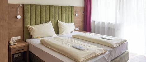 A double bed with a green upholstered headboard, two bedside tables with lamps, and a window with sheer curtains.