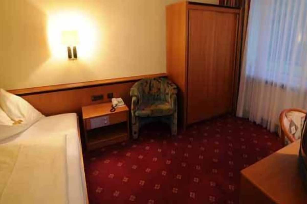 A hotel room with a bed, a chair, a desk, a wardrobe, and a lamp.