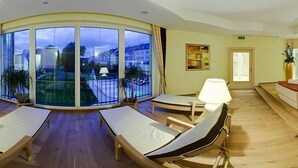 Ein sonnendurchfluteter Raum mit einer gewölbten Glaswand, zwei Lounge-Sesseln und einem kleinen Tisch mit einer Lampe.