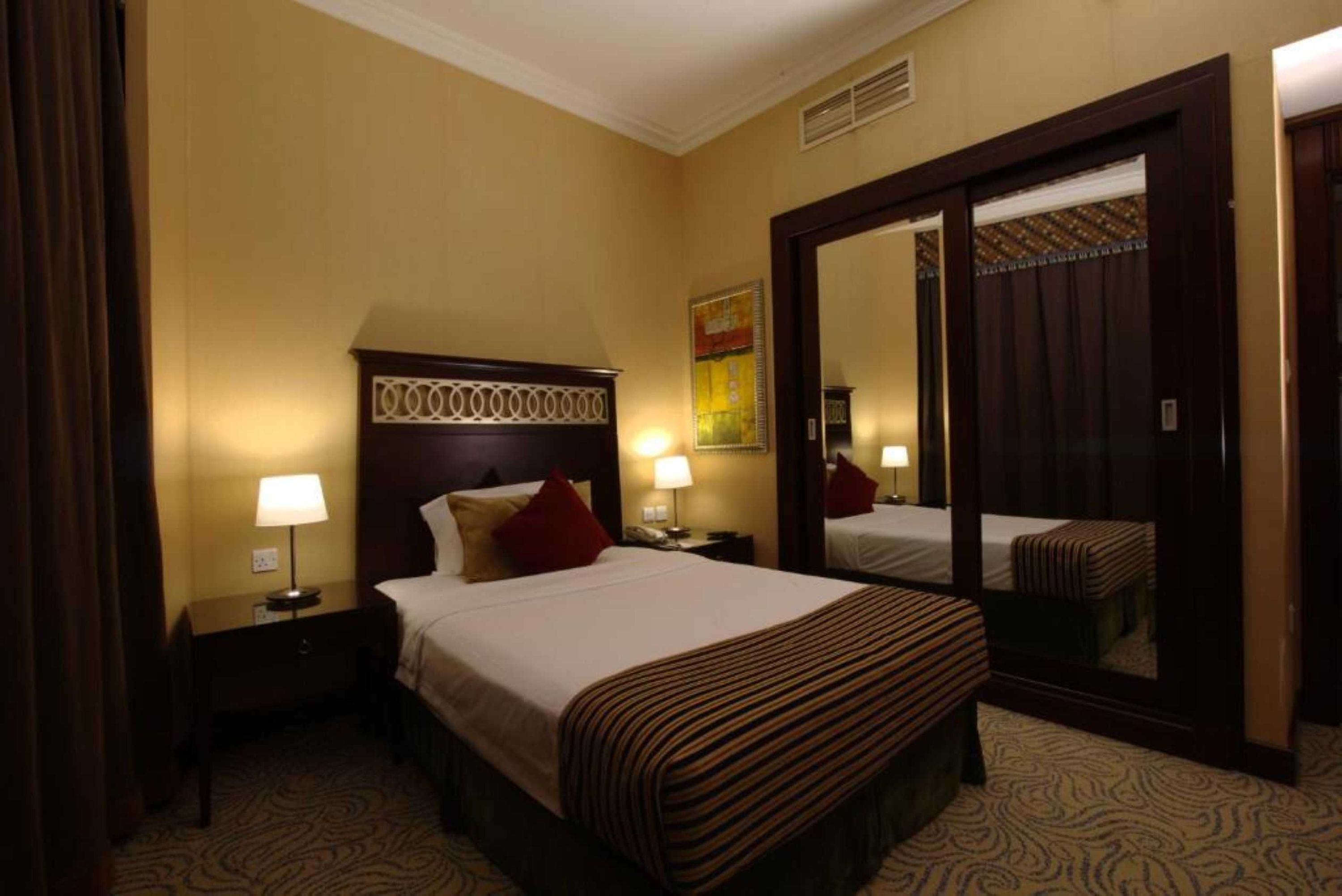 Quarto de hotel com uma cama grande, duas mesas de cabeceira com abajures, um espelho e um guarda-roupa.