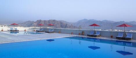 A rooftop pool with blue water, red umbrellas, and lounge chairs.
