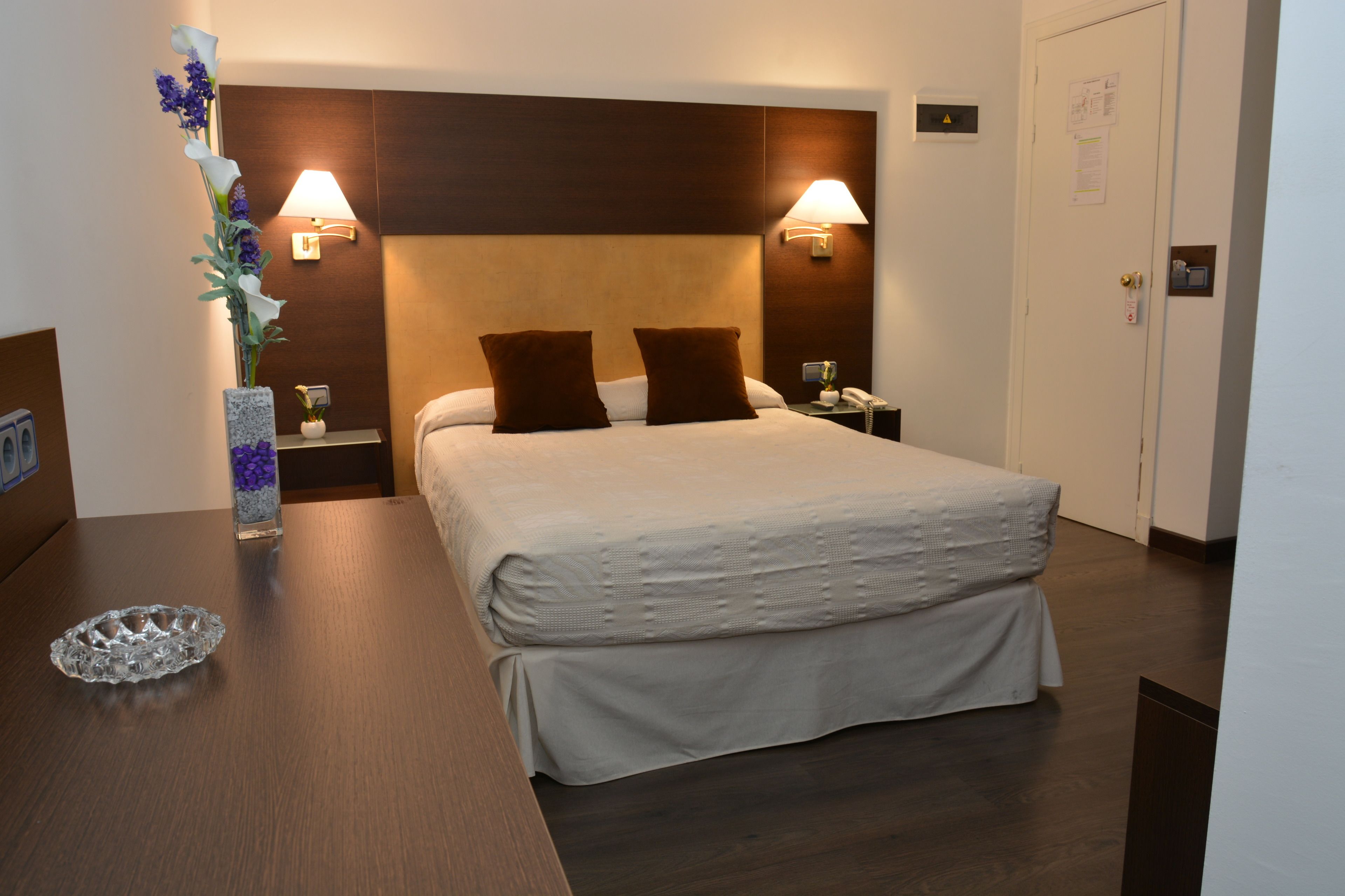 Una habitación de hotel con una cama grande, dos lámparas de noche, una mesita de noche, un jarrón con flores y un cabecero de madera.