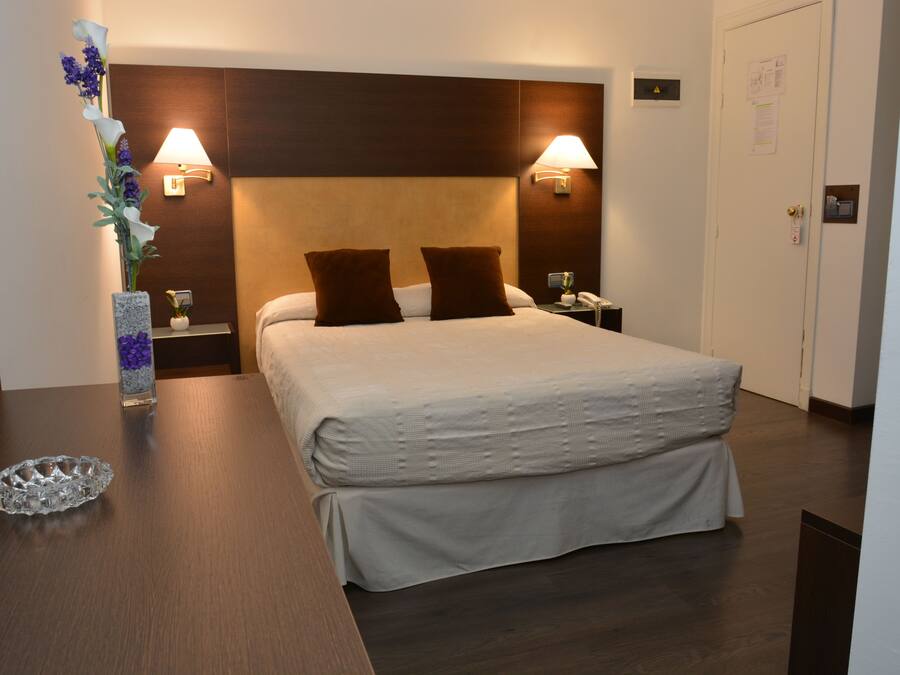 Una habitación de hotel con una cama grande, dos lámparas de noche, una mesita de noche, un jarrón con flores y un cabecero de madera.