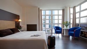 Ein Hotelzimmer mit einem großen Bett, einem Schreibtisch, zwei blauen Sesseln, einem Fenster mit Blick auf die Stadt und einer Vase mit Blumen.