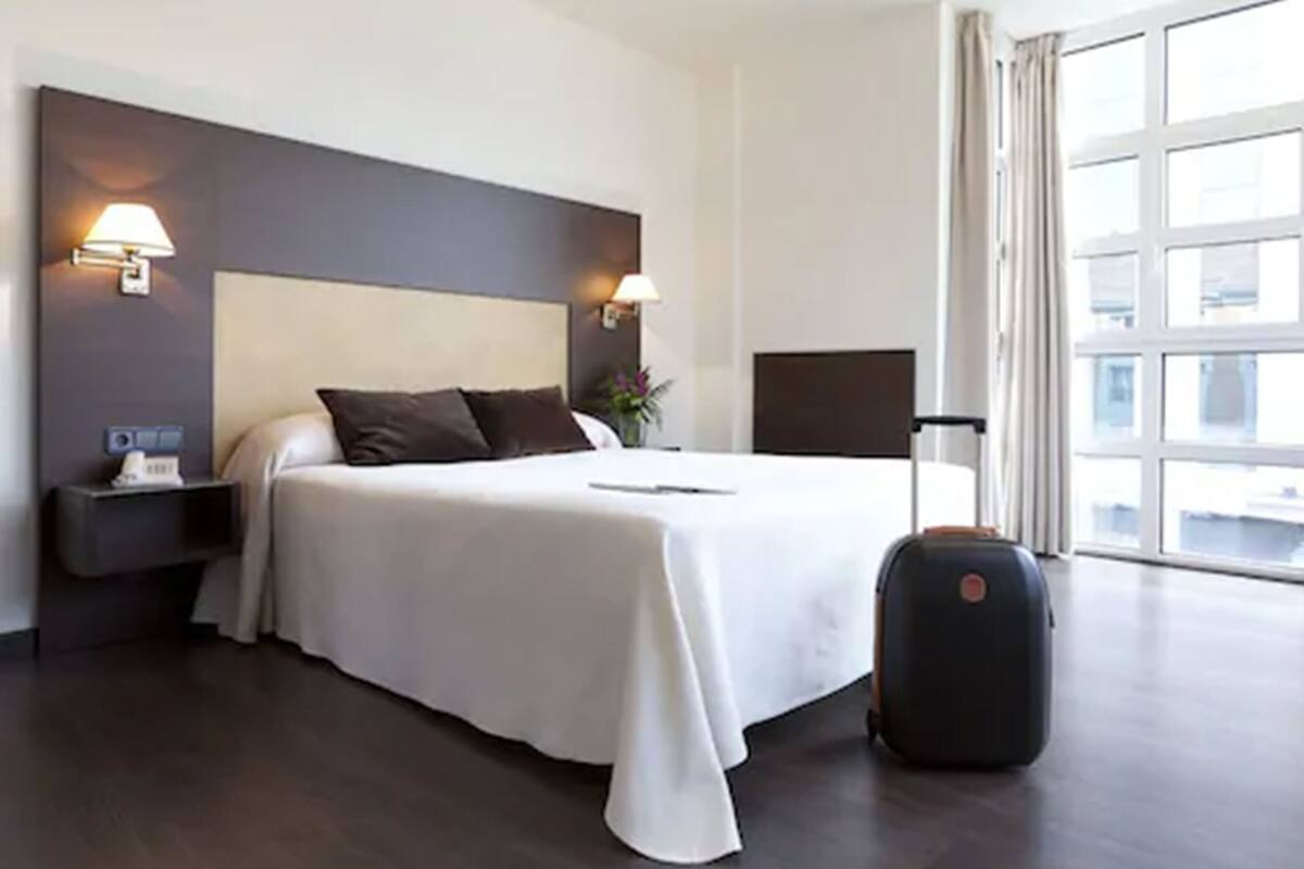 Een hotelkamer met een groot bed, twee bedlampen, een nachtkastje, een koffer en een raam met gordijnen.