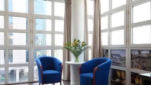 Ein Zimmer mit zwei blauen Sesseln, einem kleinen runden Tisch mit Blumenvase und großen Fenstern mit Vorhängen.