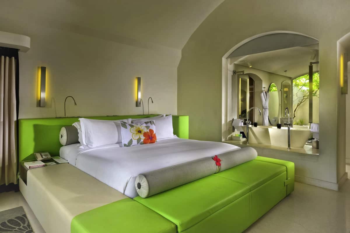 Ein Hotelzimmer mit einem großen Bett, einer grünen Polsterbank, einem Badezimmer mit einem großen Spiegel und Blick auf Grünflächen.