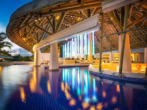 Zona de piscina con una estructura de techo de paja, asientos de descanso y el reflejo de luces en el agua.