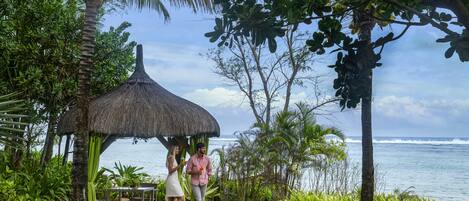 A thatched-roof structure with a table and chairs, surrounded by lush greenery and a view of the ocean.
