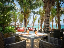 Un comedor frente a la playa con sillas y mesas de mimbre, palmeras y vistas al océano.