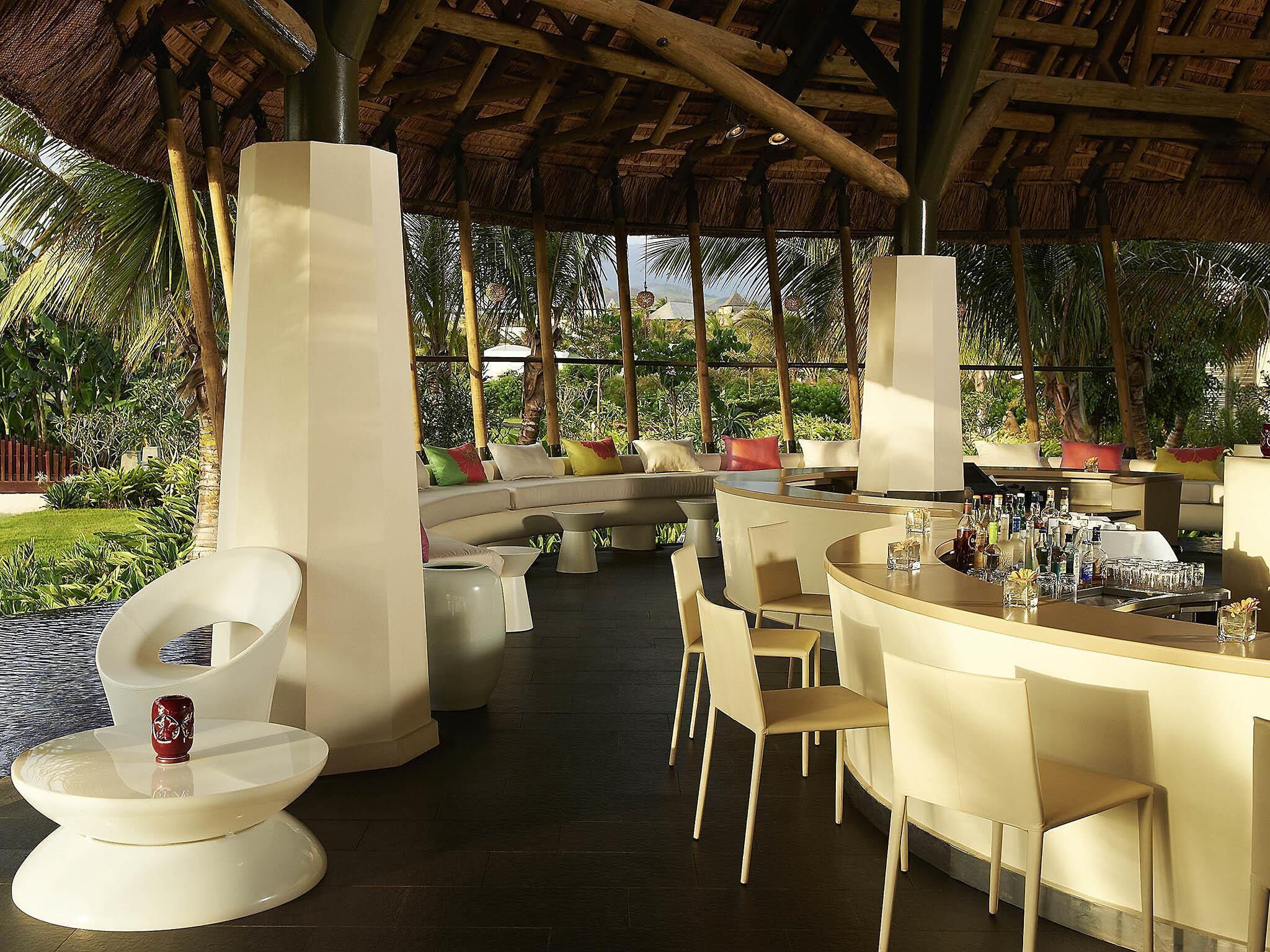 Un espacio lounge moderno al aire libre, con una disposición de asientos curva, una barra con varias botellas y una estructura de techo de paja.