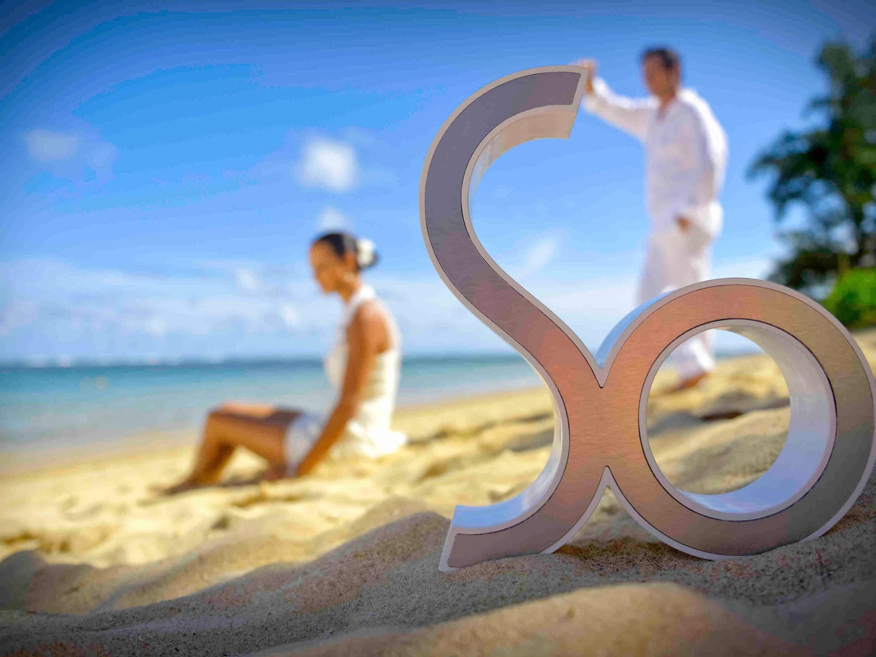 Escena de playa con un gran símbolo de infinito en la arena, una persona sentada y otra de pie al fondo.
