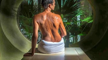 Una persona sentada en una bañera circular, con una toalla alrededor de la cintura, rodeada de plantas verdes.