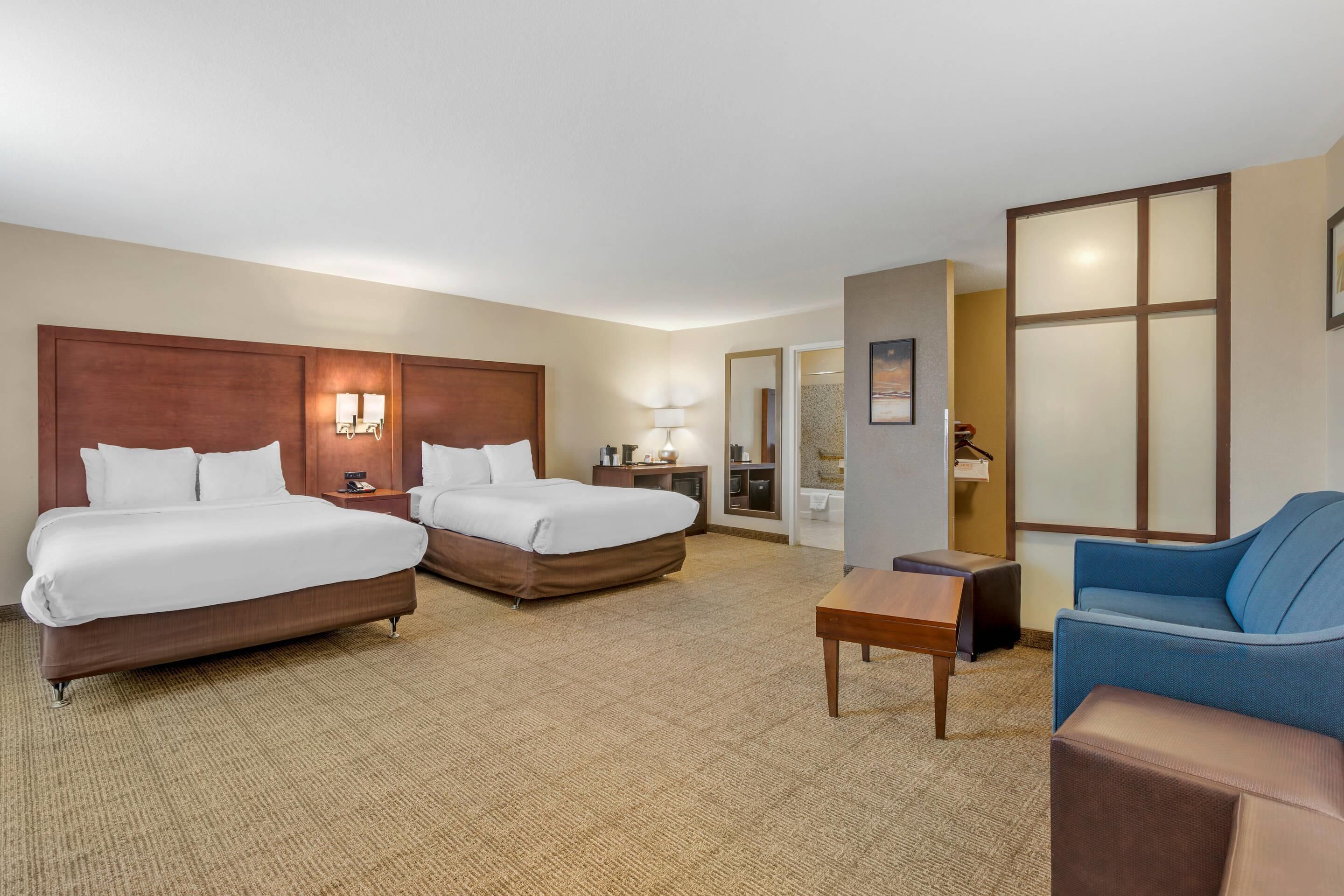 Habitación de hotel con dos camas, un sofá, una mesita y un baño visible a través de una puerta de cristal.