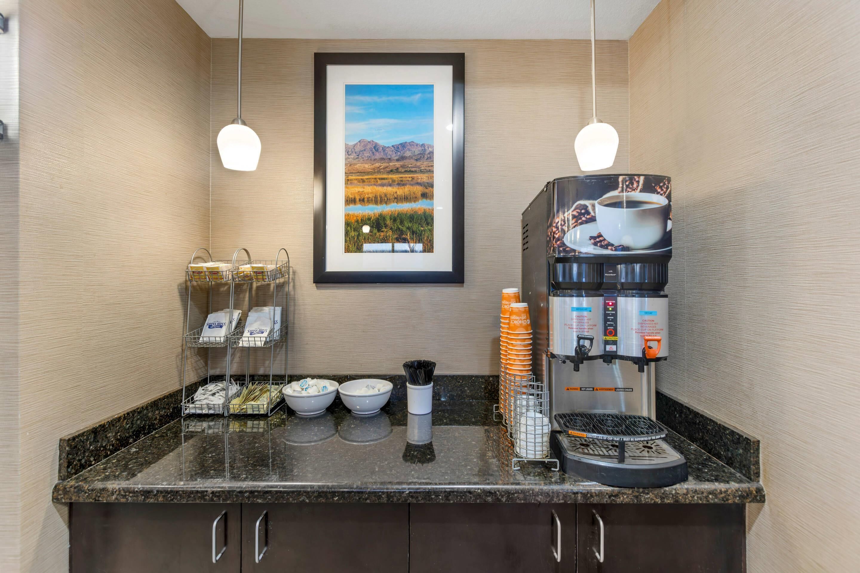 Cozinha compacta de hotel com máquina de café, tigelas e uma imagem emoldurada de uma paisagem.