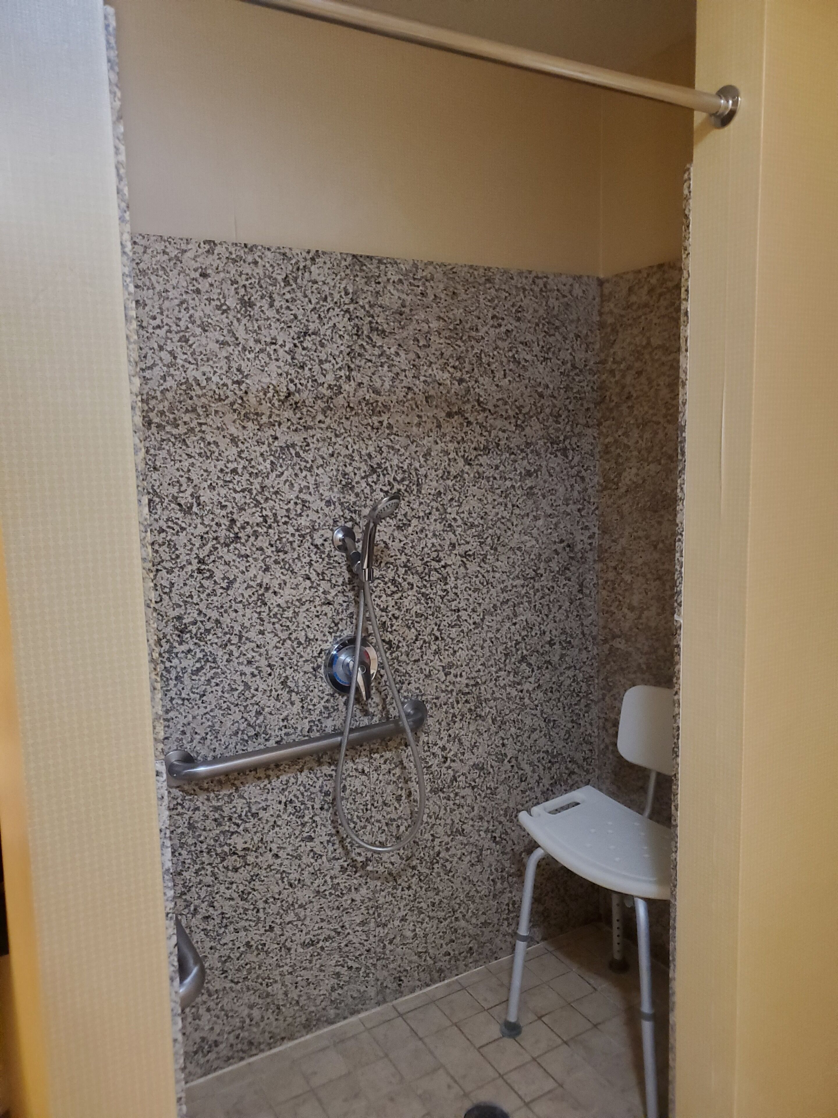A bathroom with a granite wall, a shower with a handheld showerhead, a white chair, and a handrail.