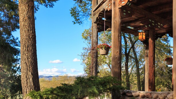 A wooden gazebo with hanging flower baskets and lanterns, surrounded by trees and a stone wall.