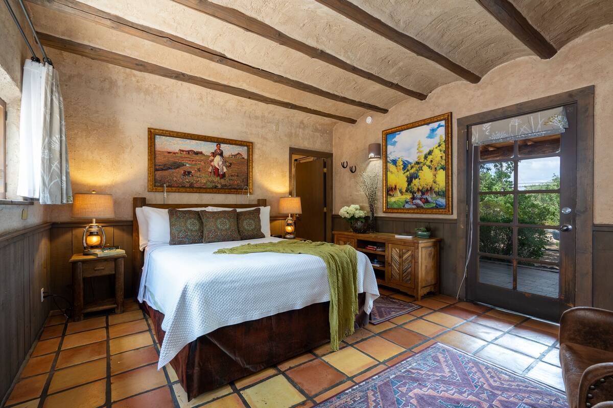 A bedroom with a large bed, wooden headboard, two bedside lamps, a nightstand, a dresser, a door leading outside, and two paintings on the walls.