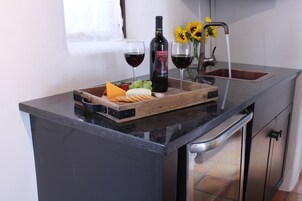 A kitchen counter with a wooden board holding cheese, grapes, and crackers, a bottle of wine, and two glasses of red wine.