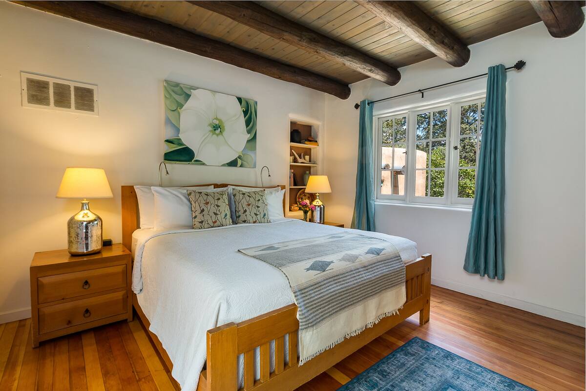 A bedroom with a wooden bed, two bedside tables with lamps, a large floral painting, and a window with curtains.