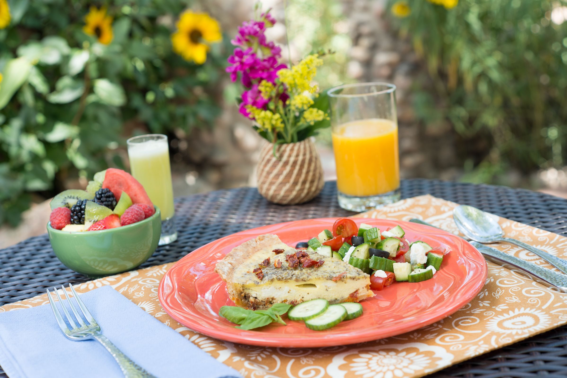 A meal with a slice of quiche, salad, and a bowl of fruit on a table with a woven placemat.