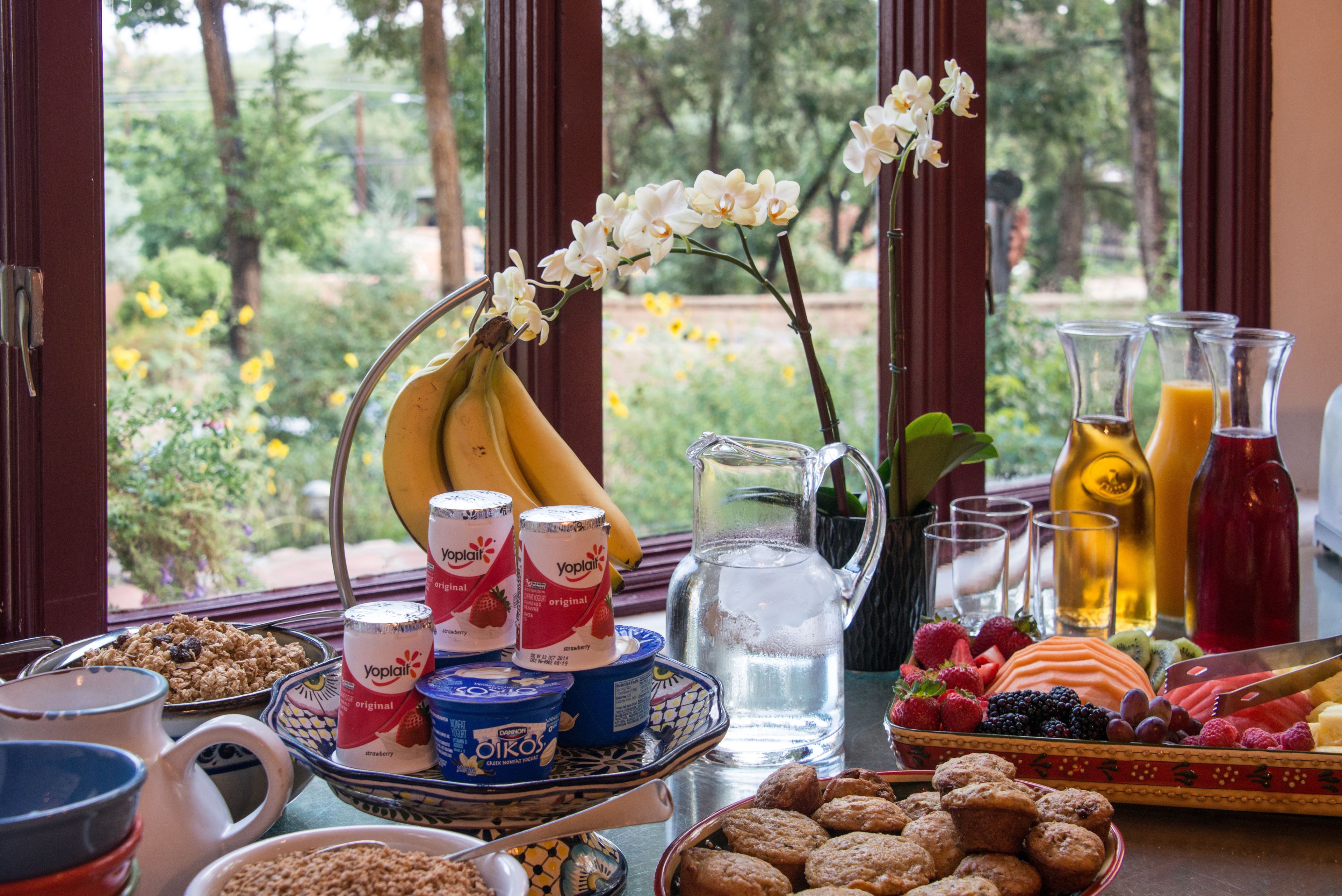 A breakfast table with various food items, including fruits, pastries, and beverages, arranged on a table near a window with a view of greenery.