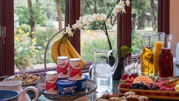 A breakfast table with various food items, including fruits, pastries, and beverages, arranged on a table near a window with a view of greenery.