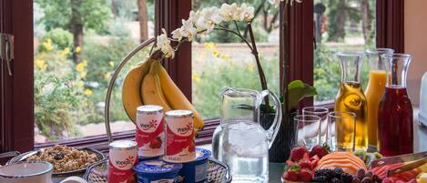A breakfast table with various food items, including fruits, pastries, and beverages, arranged on a table near a window with a view of greenery.