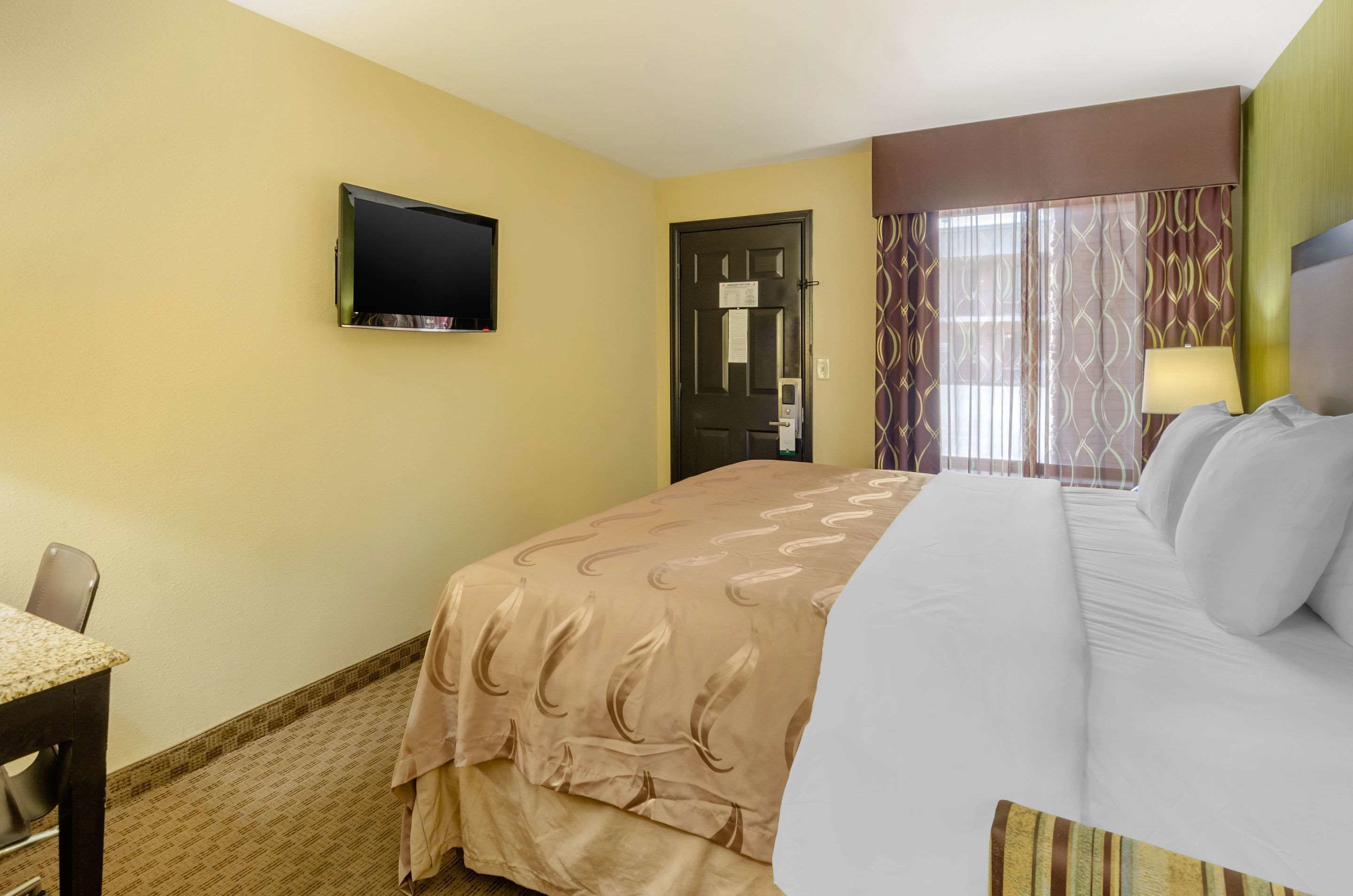 Una habitación de hotel con cama, televisor montado en la pared, una puerta y una ventana con cortinas.